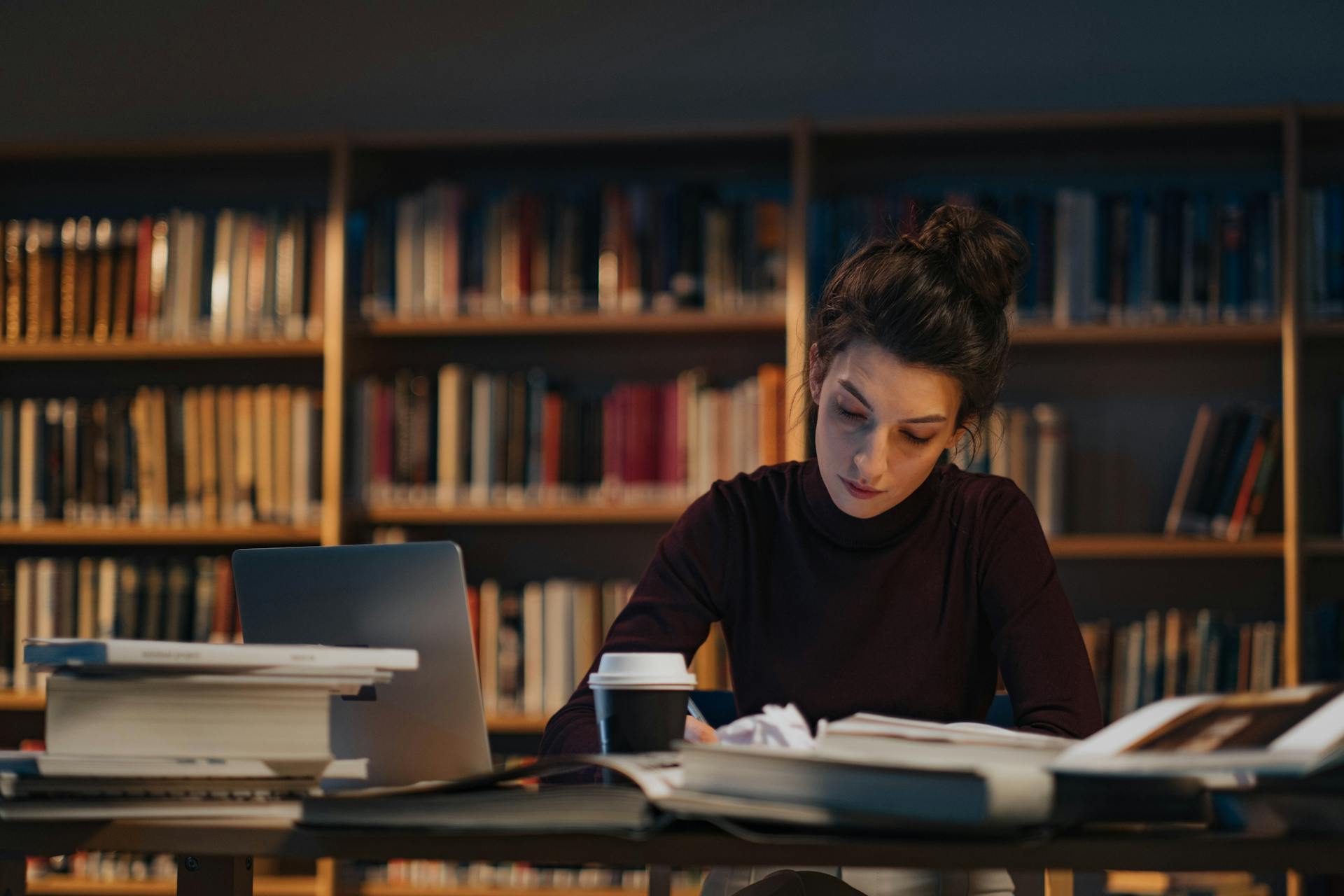 A researcher in a library at evening — laptop open, books stacked beside her, mid-thought over an open text.